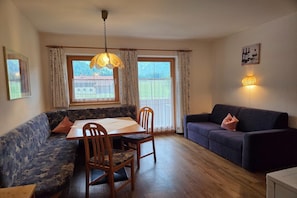A cozy living room with a dining table, wooden chairs, a sofa, and a couch. There are two windows with curtains, a hanging light fixture, and a wall-mounted picture. - Holiday apartment near Lake Achensee (Achenkirch)