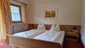 Habitación de hotel con cama de madera, ropa de cama blanca, mesita de noche con lámpara, radiador y ventana con cortinas.