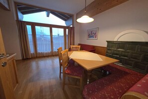 Ein Essbereich mit Holztisch, Stühlen und Bänken. Ein Fenster mit Vorhängen, eine Wandleuchte und ein Wandbild sind zu sehen.