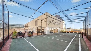 Uma quadra de tênis com rede, cercada por uma grade, e um edifício ao fundo.