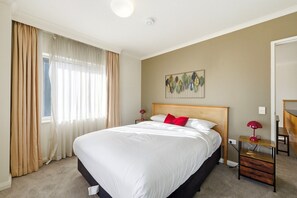 A hotel room with a large bed, a bedside table with a lamp, a wall art piece, and a window with curtains. - Stylish 2-Bed Stay with Pool & Parking (City)
