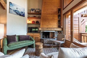 Ein gemütliches Wohnzimmer mit Kamin, Holzregalen, einem Fernseher und einem grünen Sofa.