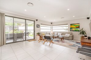 A spacious living room with a sliding glass door leading to a patio, a sofa set, a coffee table, and a painting on the wall. - Charming 4-Bed Getaway with Pool & Garden Oasis (Curtin)