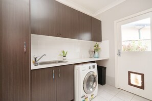A compact kitchen with a built-in washing machine, a sink, and a small shelf with decorative items. - Charming 4-Bed Getaway with Pool & Garden Oasis (Curtin)