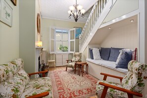 A cozy living room with floral-patterned furniture, a red patterned rug, a white bench with blue cushions, and a staircase with white railings. - Tranquil Cottage | Lovely Street, Walk to Leura (Leura)