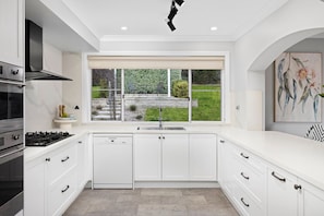 A modern kitchen with white cabinetry, a built-in dishwasher, a gas stove, and a window with a view of a garden. - Carinya Centennial | Bowral Spa Retreat (Bowral)