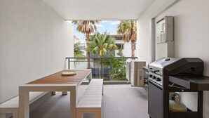 A balcony with a table and grill, surrounded by white walls and a view of palm trees and buildings. - Beach & Lake Narrabeen Apartment with Outdoor BBQ (Narrabeen)
