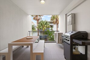 Ein Balkon mit Tisch und Grill, umgeben von weißen Wänden und Blick auf Palmen und Gebäude.