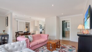 Un salon moderne avec un canapé rose, un fauteuil gris et une table basse en bois. On aperçoit à l’arrière-plan un coin repas avec une table et des chaises.