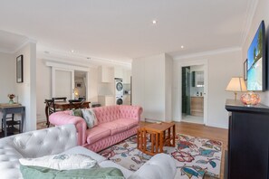 Ein modernes Wohnzimmer mit einem rosafarbenen Sofa, einem grauen Sessel und einem Holztisch. Im Hintergrund ist eine Essecke mit Tisch und Stühlen zu sehen.