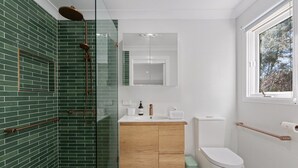 Une salle de bain dotée d’une douche à parois de verre, d’un meuble-lavabo en bois, de toilettes et d’une fenêtre donnant sur la forêt.