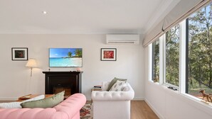Un salon avec un canapé rose, un fauteuil blanc, une cheminée et une grande fenêtre donnant sur la forêt.