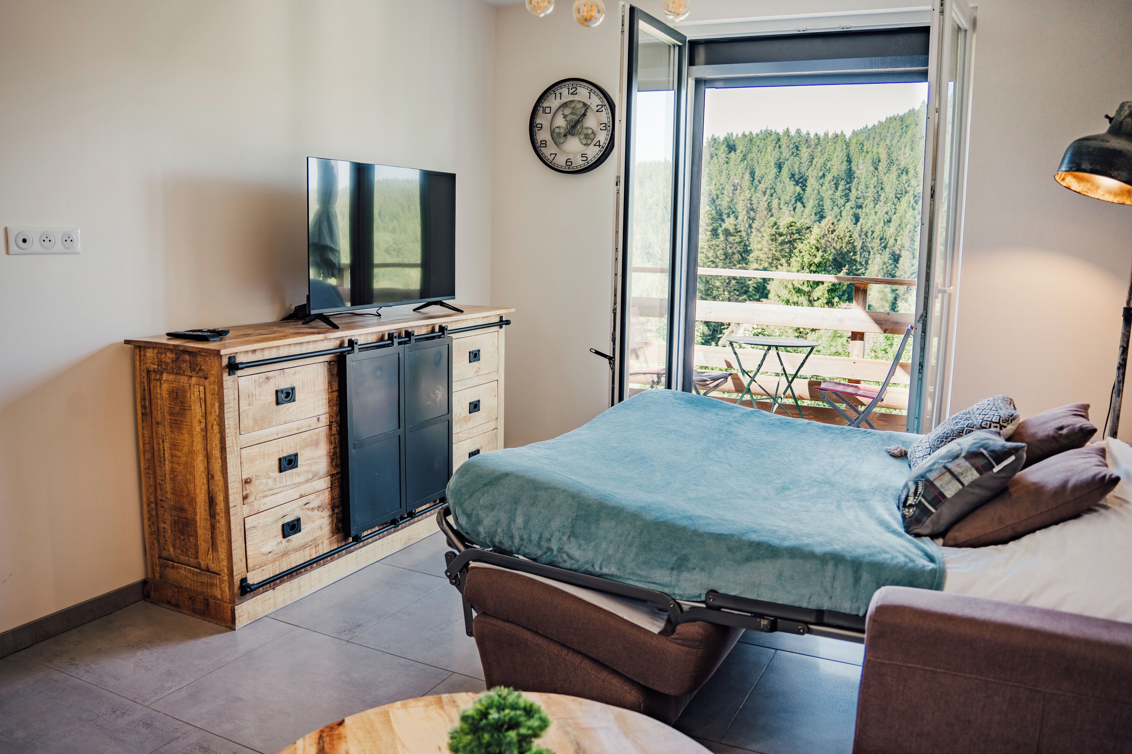 Een slaapkamer met een bed, een televisie, een dressoir en een balkon met uitzicht op bomen.