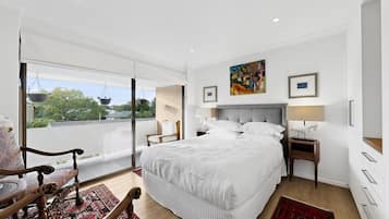 A modern bedroom with a large bed, a bedside table, a chair, a window with a view, and a painting on the wall.