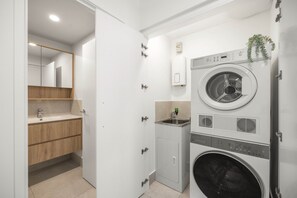 A compact laundry room with a front-loading washing machine, a small sink, and a mirror. - Poolside 2-Bed Apartment at Lake Kawana (Birtinya)