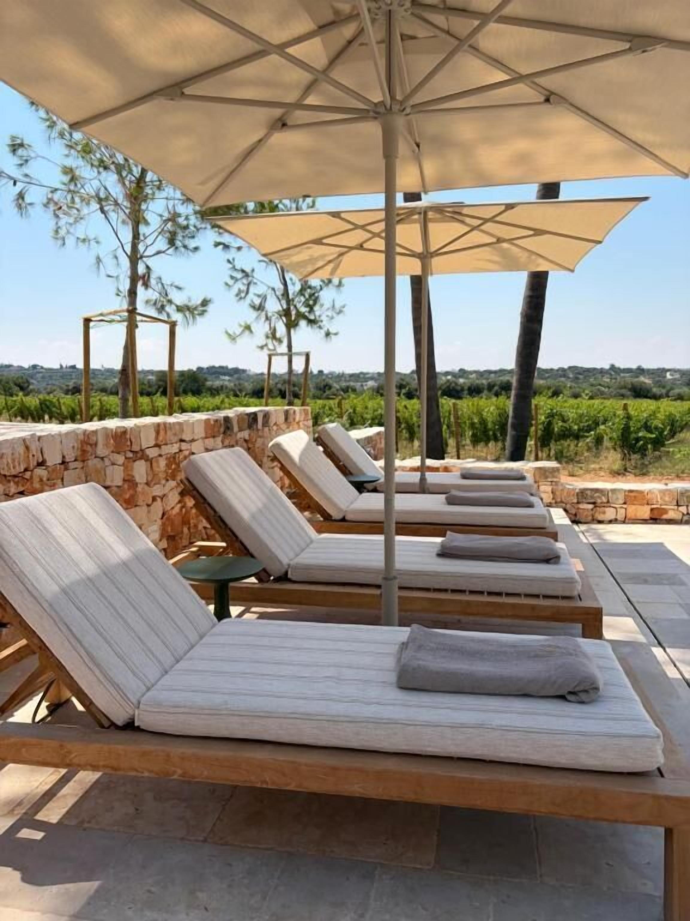 A row of lounge chairs with cushions, a large umbrella, and a stone wall backdrop.