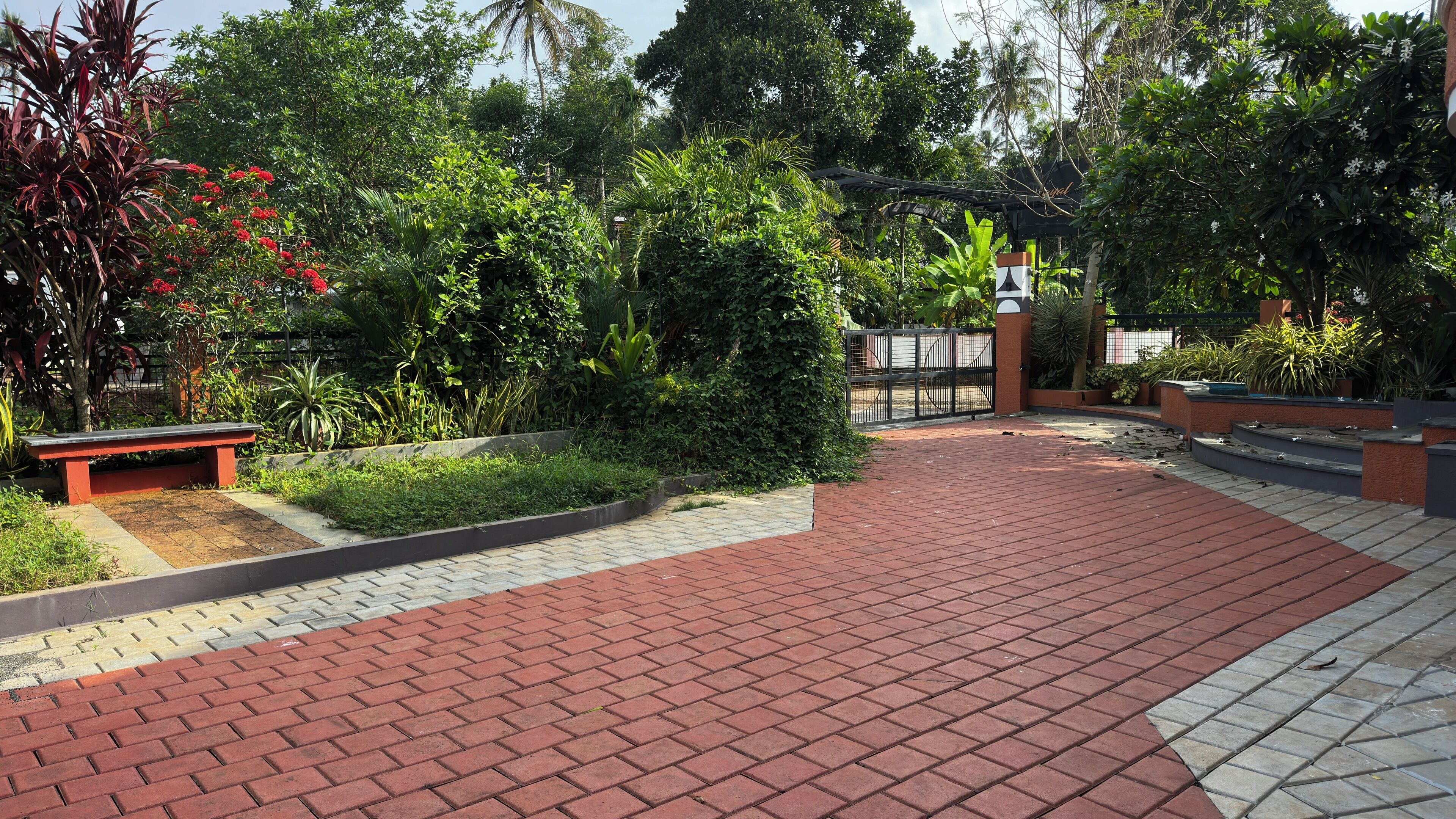A paved walkway with a bench, surrounded by lush greenery and a gated entrance.