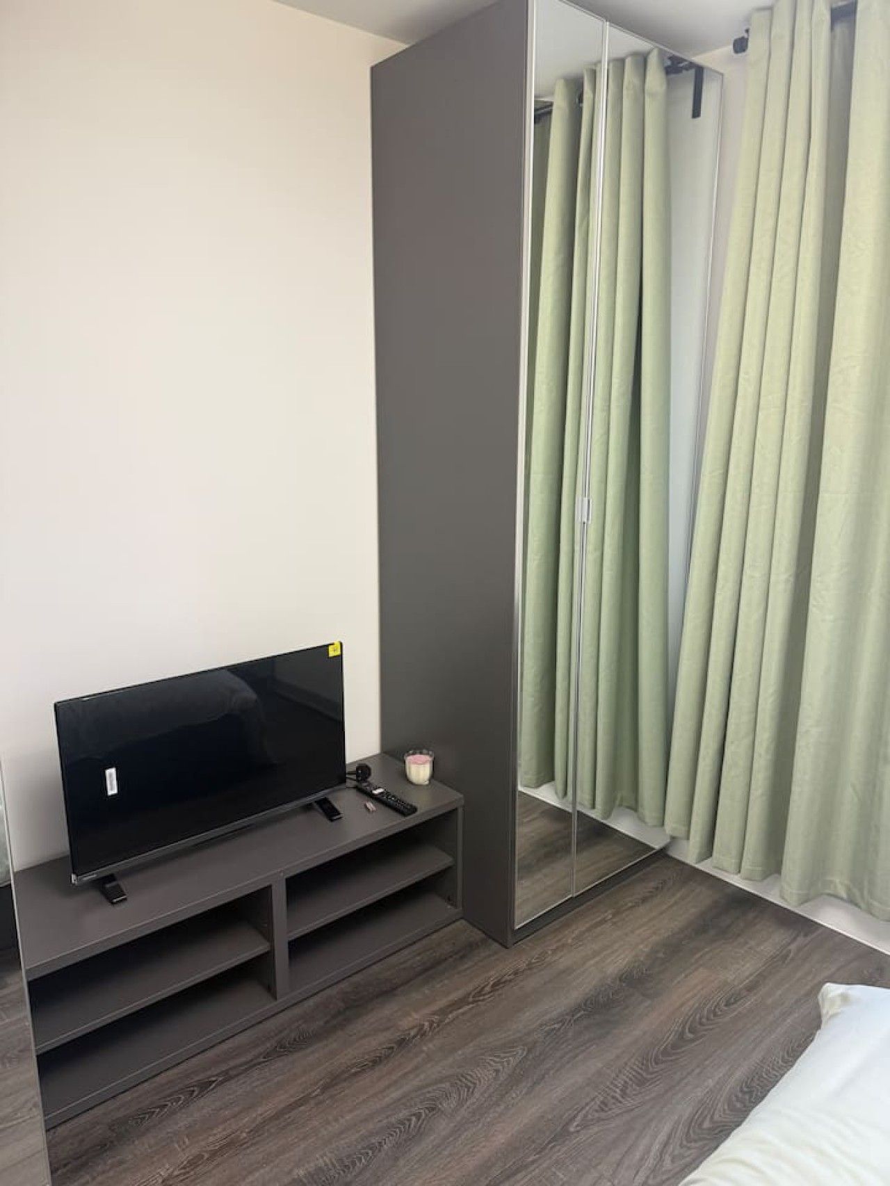 A modern bedroom with a flat-screen TV on a dark wooden stand, a small bedside table with a remote and a candle, and a large mirror reflecting a green curtain.
