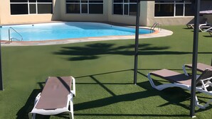 A pool area with artificial grass, lounge chairs, and a building with large windows. - Vacation Home 'Casa Del Mar' with Shared Pool, Private Garden and Wi-Fi (Costa Calma)