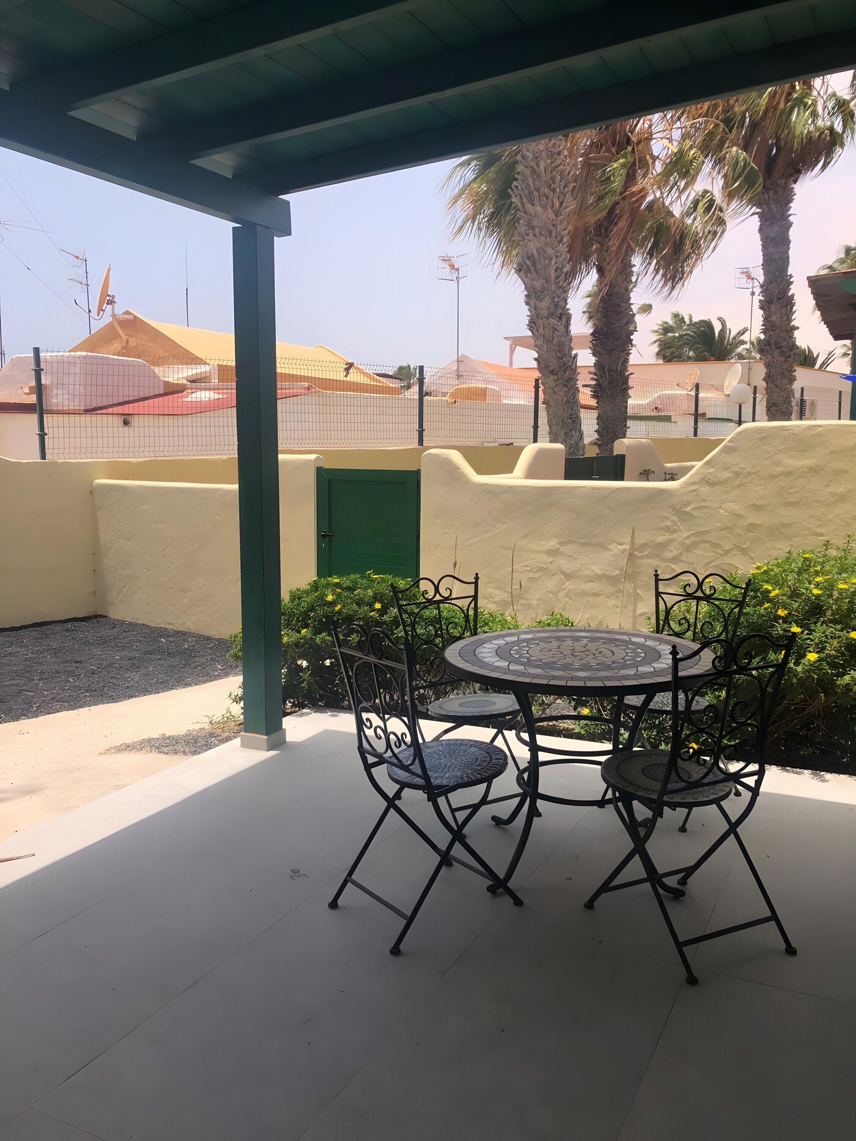 A covered outdoor seating area with a table and chairs, a green door, and a view of a fenced sports facility.