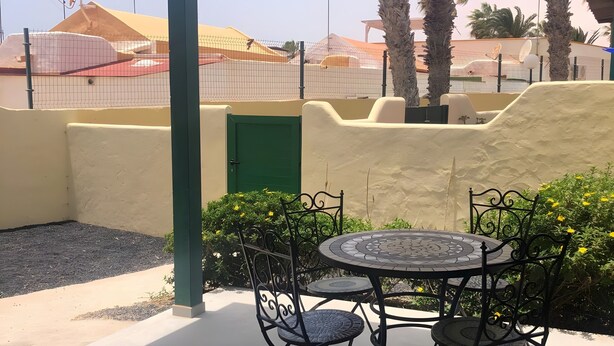 A covered outdoor seating area with a table and chairs, a green door, and a view of a fenced sports facility. - Vacation Home 'Casa Del Mar' with Shared Pool, Private Garden and Wi-Fi (Costa Calma)