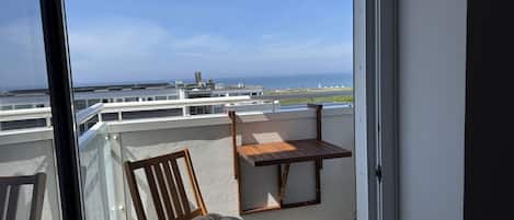 A balcony with a wooden chair and a small table, overlooking a coastal view with buildings in the distance.