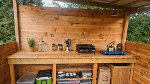 Uma cozinha externa de madeira com mesa integrada, recipientes de armazenamento e utensílios de cozinha.