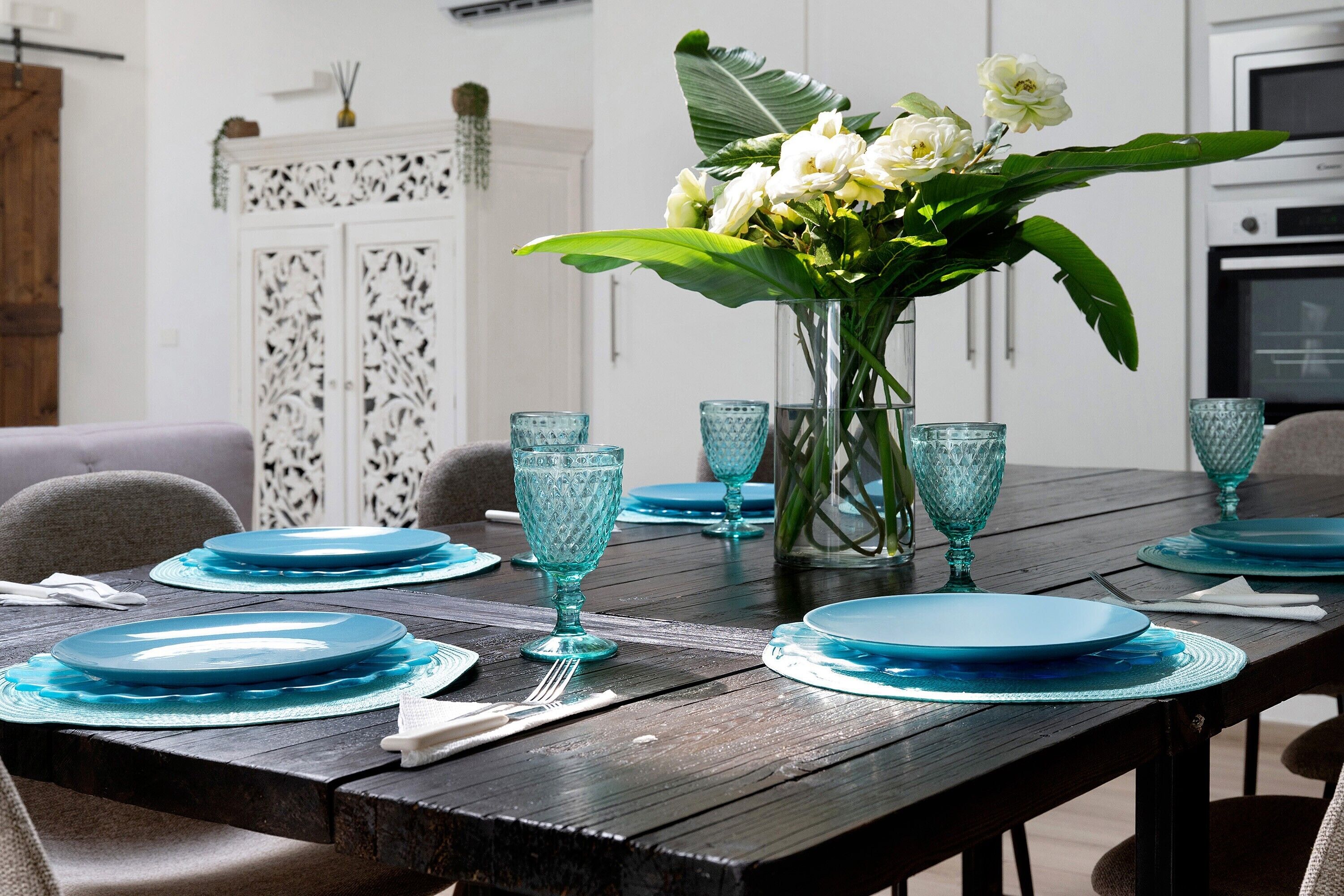 Ein Esstisch, gedeckt mit blauem Glas und Geschirr, eine Vase mit Blumen und ein weißer Schrank im Hintergrund.