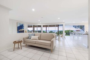 A modern living room with a beige sofa, a small wooden side table, and a view of the outdoors through large windows.
