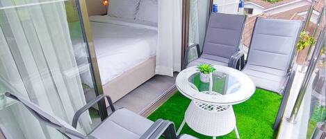 A balcony with a bed, lounge chairs, a small table with a plant, and a view of buildings.