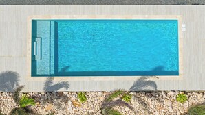 Une piscine rectangulaire avec une terrasse carrelée, entourée d’une bordure de galets et de plantes tropicales.