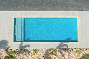 A rectangular swimming pool with a tiled deck, surrounded by a pebble border and palm plants. - Moyo Apartment. 2 bedrooms & 2 bathrooms. 15-minute drive to Jan Thiel & Mambo (Willemstad)