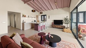 Un salon moderne avec un canapé, une table basse et un téléviseur. Il y a également un coin cuisine avec un bar et une table à manger. La pièce est dotée d’un haut plafond avec des poutres en bois.
