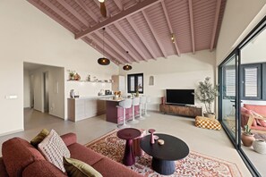 A modern living room with a sofa, a coffee table, and a TV. There is a kitchen area with a bar and a dining table. The room has a high ceiling with wooden beams. - Moyo Apartment. 2 bedrooms & 2 bathrooms. 15-minute drive to Jan Thiel & Mambo (Willemstad)