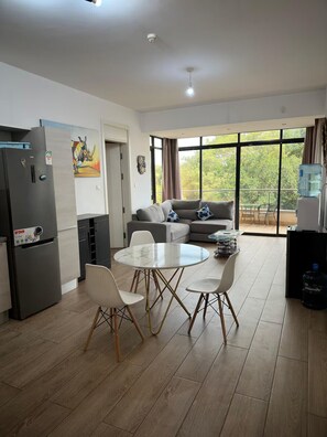 A modern kitchen with a refrigerator, dining table, and chairs. There is a sofa and a large window with a view of trees. - 2-bedroom apartment in adorable Nairobi with fitness room access (Nairobi)