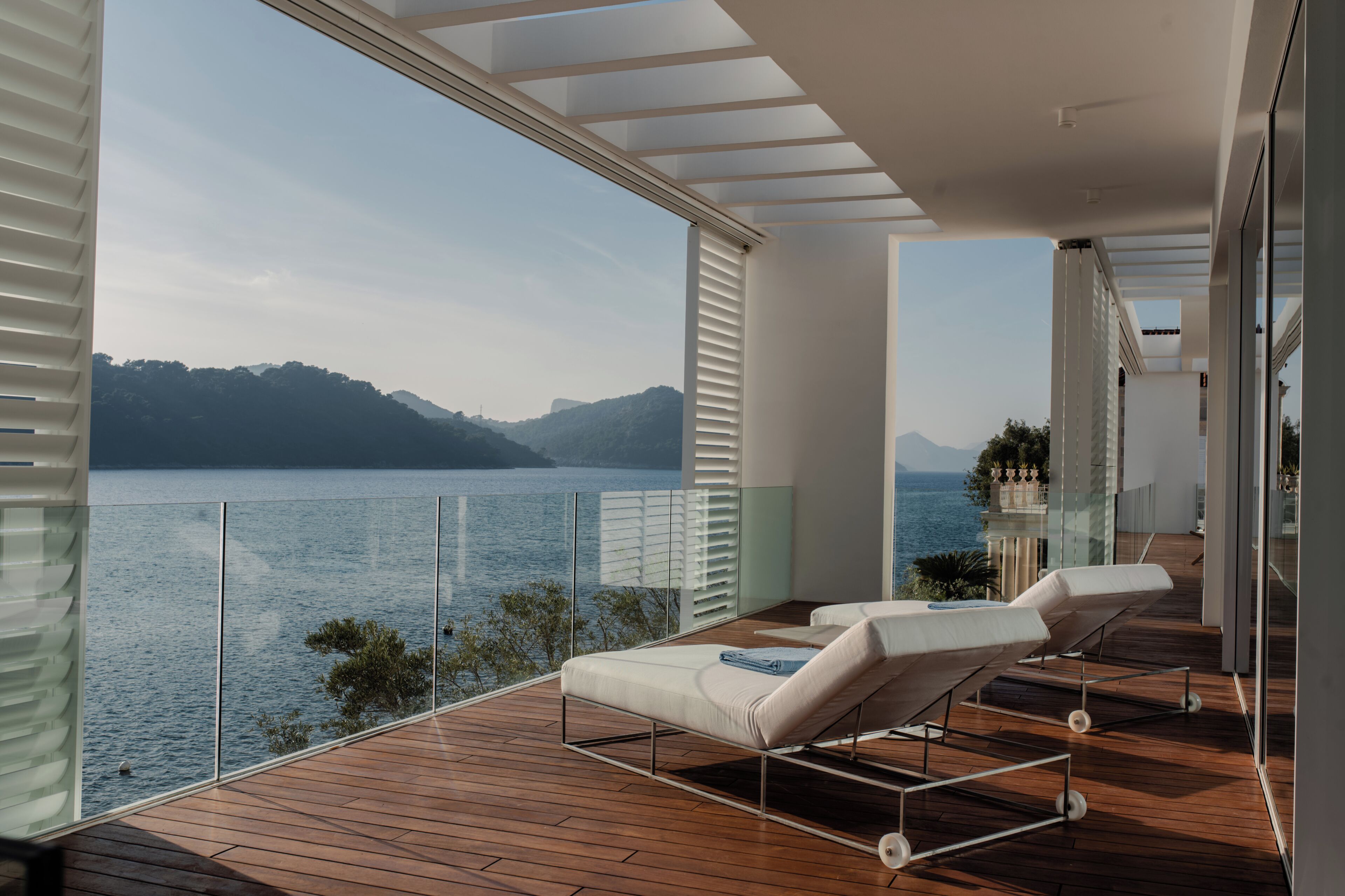 Un balcon moderne avec un plancher en bois, deux fauteuils blancs et une balustrade en verre donnant sur un lac.