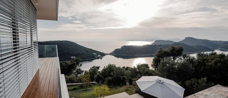 A balcony with a view of a lake and mountains, a wooden deck, and outdoor umbrellas.