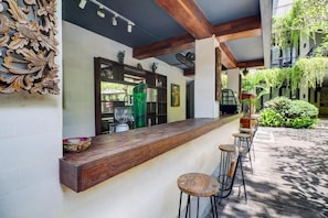A bar area with a wooden counter and stools, a decorative wall panel, and a view of a lush garden.