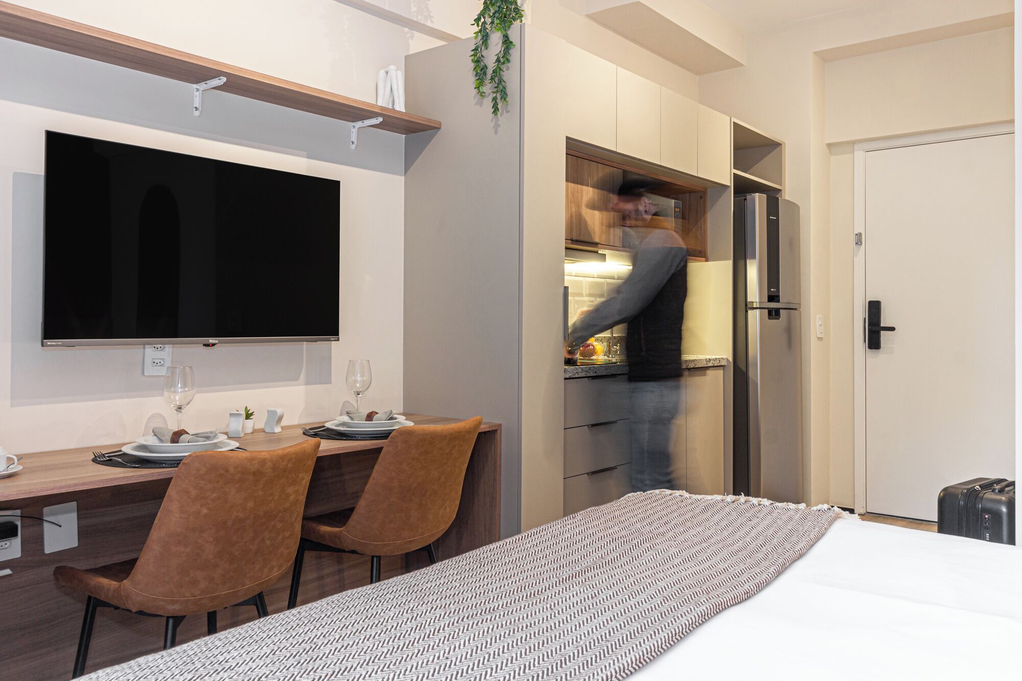 A modern hotel room with a dining area, a flat-screen TV, a bed, and a kitchenette.