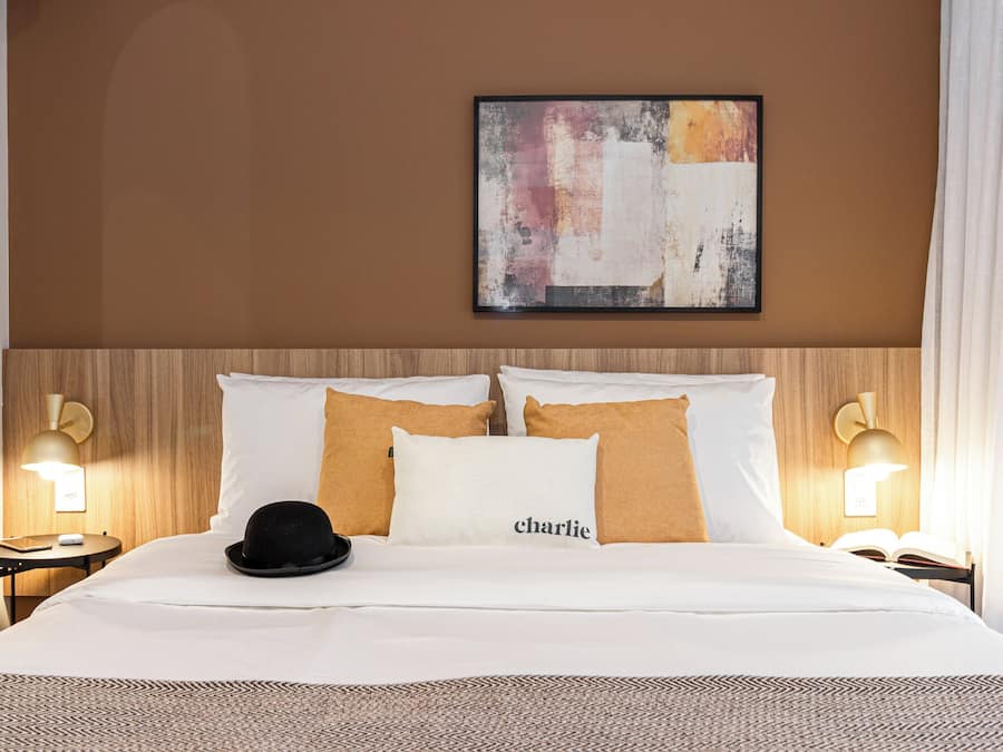 Une chambre d’hôtel avec un lit, deux lampes de chevet, un chapeau posé sur le lit et un tableau accroché au mur.