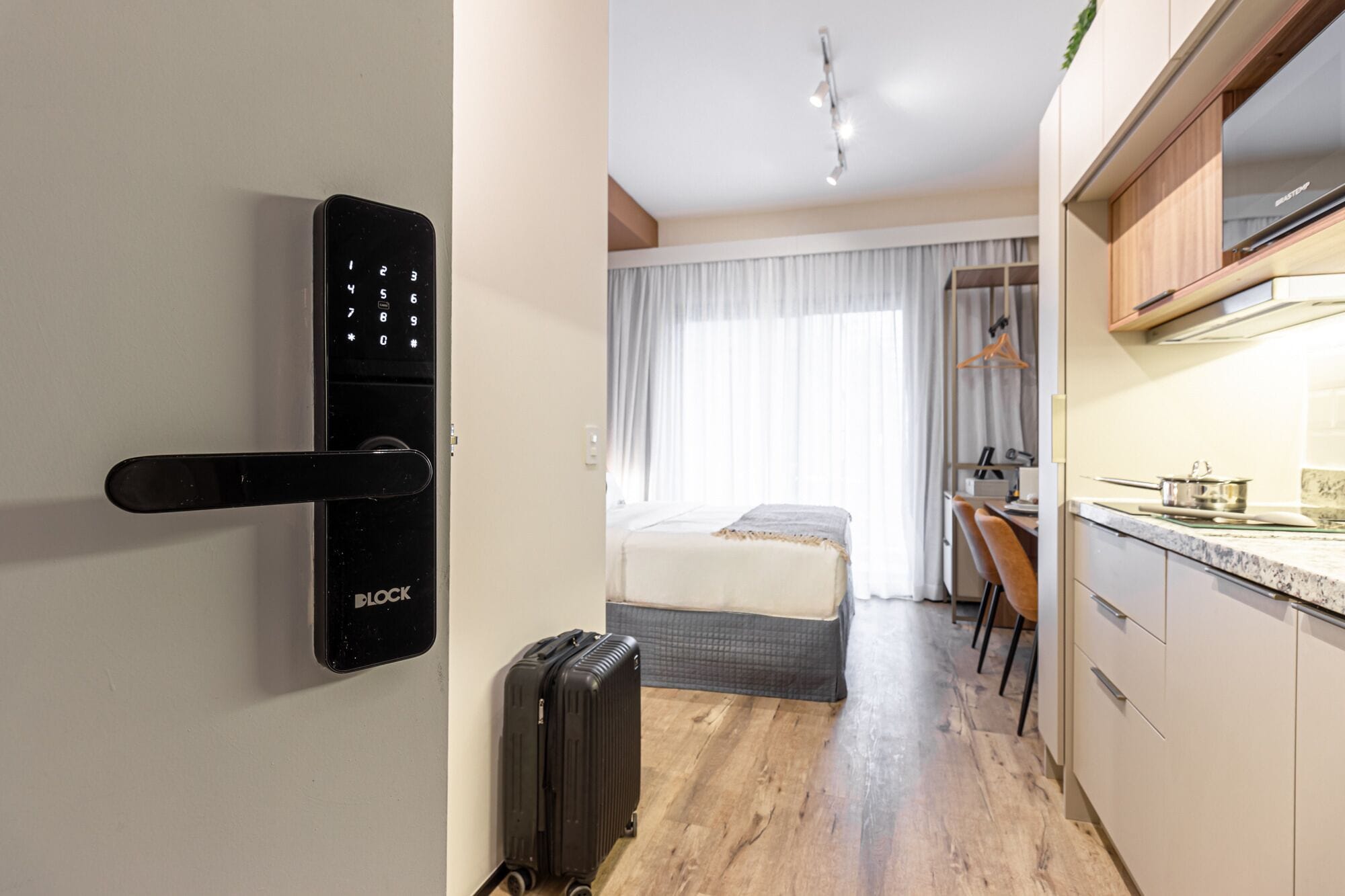 A modern hotel room with a digital lock, a bed, a small kitchenette, and a suitcase.