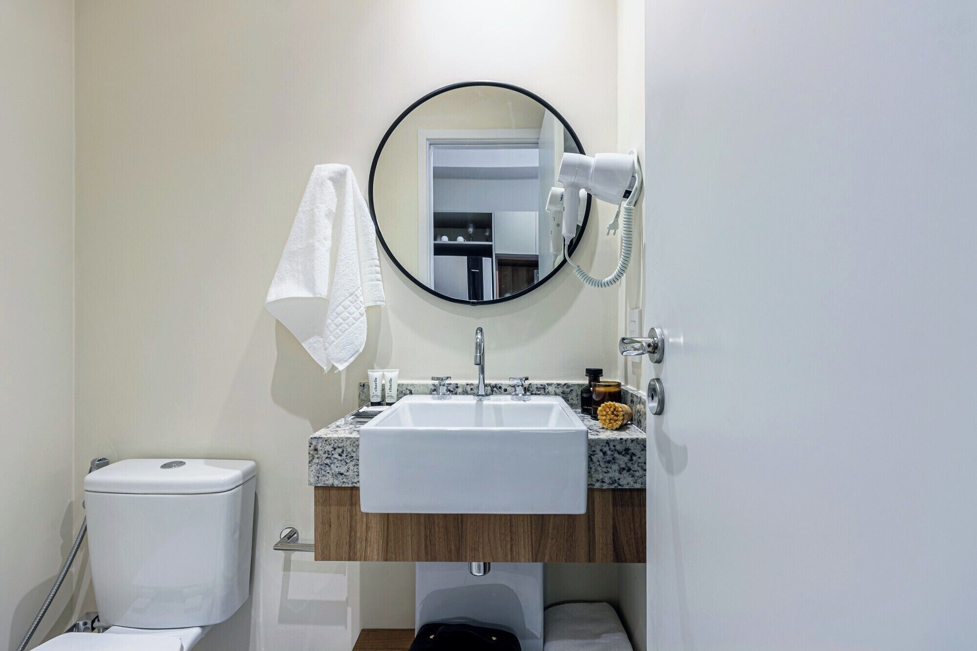 Une salle de bain avec un lavabo blanc, un miroir rond, un sèche-cheveux et des toilettes.