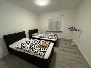 Ein kleines, sauberes Zimmer mit zwei Betten, einem Fenster mit durchsichtigen Vorhängen und einem Wäscheständer.