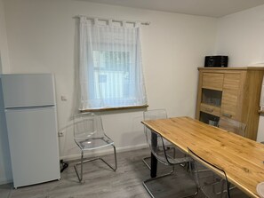 A small kitchen with a refrigerator, a wooden table, and a window with sheer curtains.