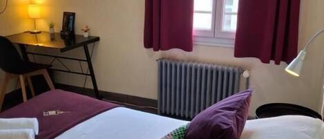 Une chambre d’hôtel avec un lit, un bureau, une chaise, un radiateur et une fenêtre avec des rideaux.