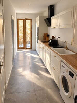 A modern kitchen with a built-in washer, a stone wall, and a large sliding glass door leading to a patio. - Rose des Orpellières – Terrace, A/C and shared garage (Sérignan)