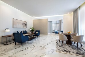 Quarto de hotel moderno com piso de mármore, um sofá, uma mesa de jantar com cadeiras, uma mesa de centro e uma janela grande com cortinas.