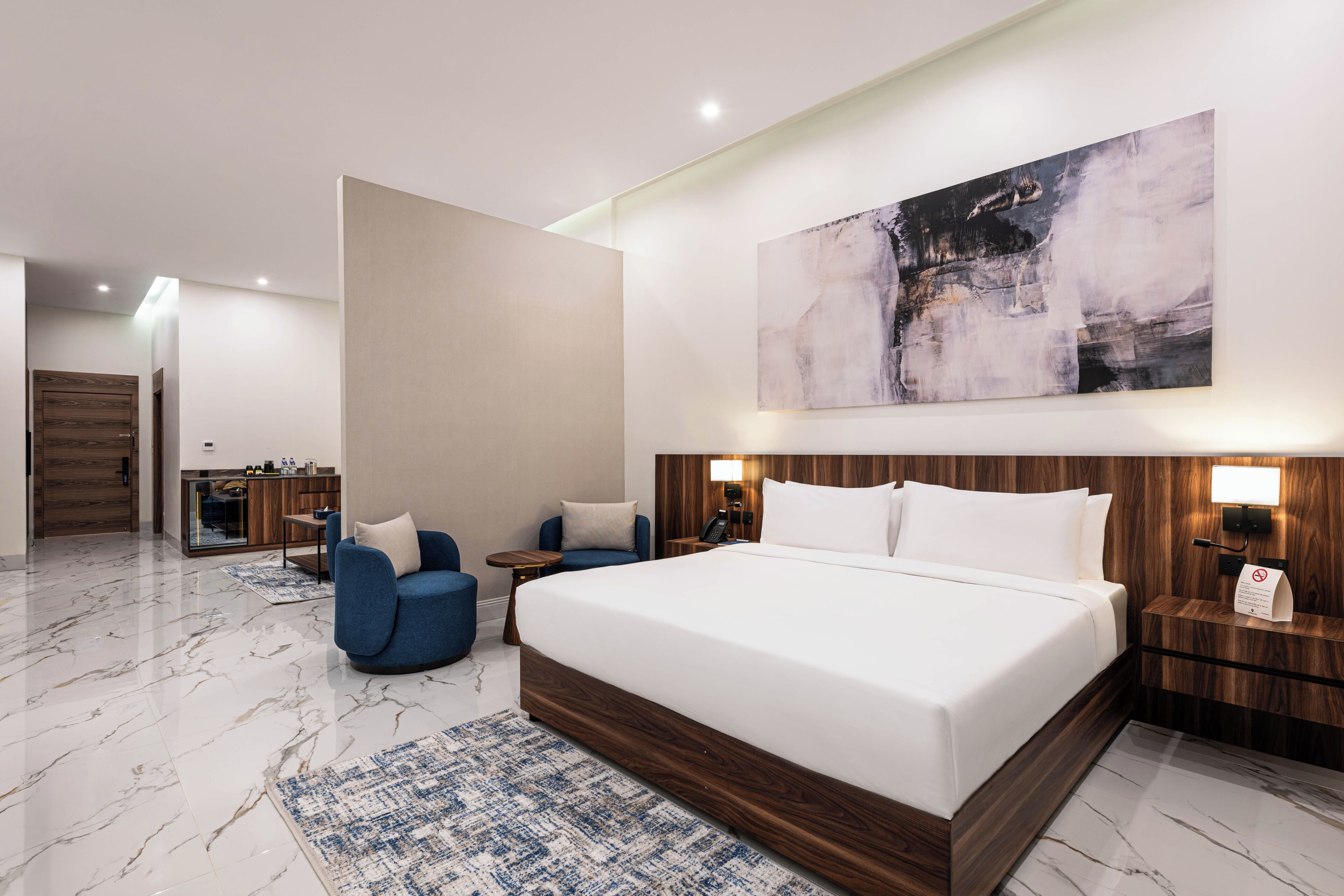 A modern hotel room with a large bed, two armchairs, a small table, a desk with a phone, and a painting on the wall.