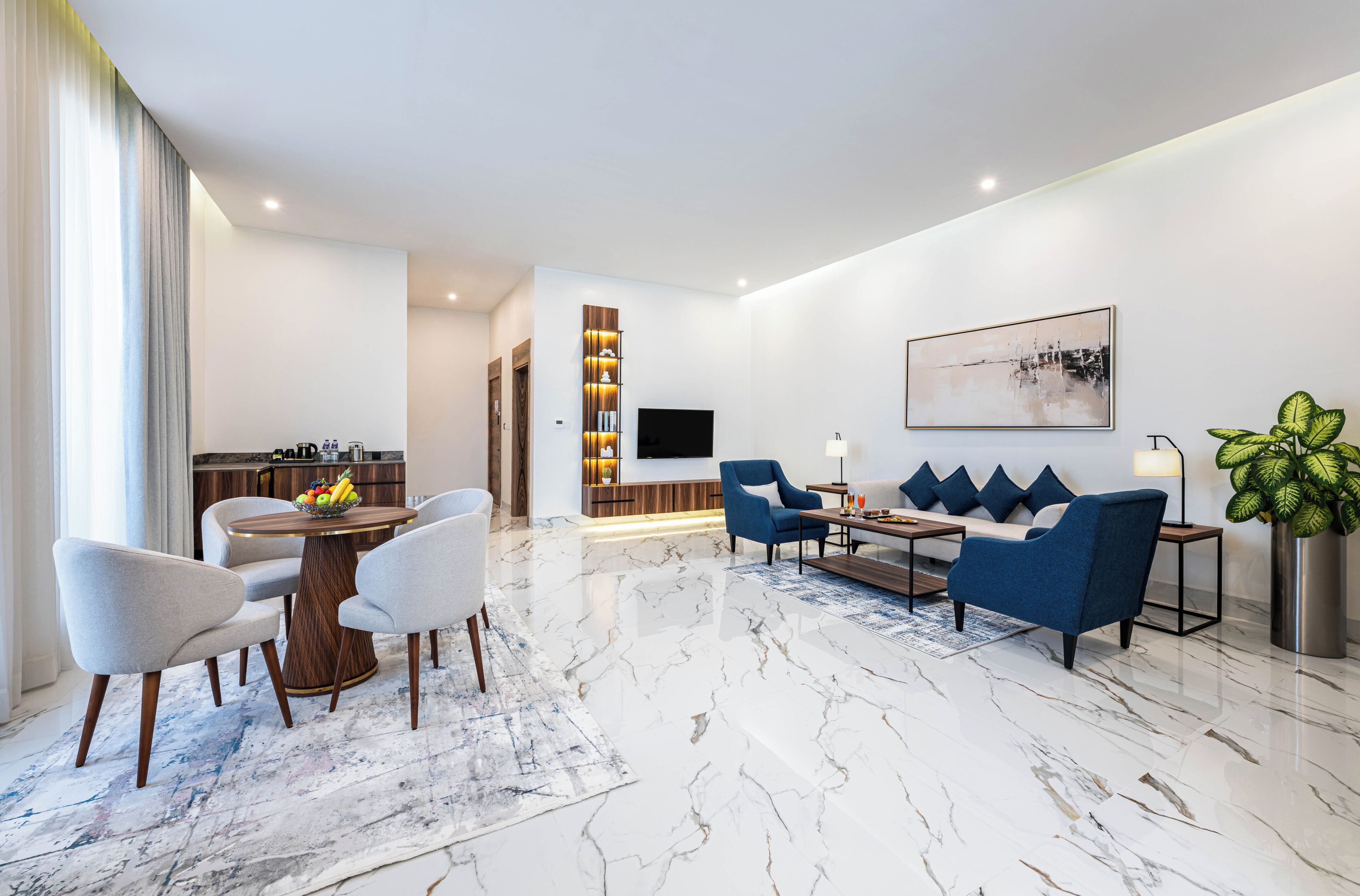 Una sala de estar moderna con piso de mármol, un comedor con frutero, y una zona de estar con sofás azules y una mesa de centro.