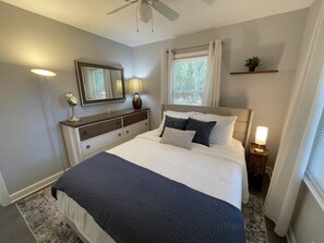 A bedroom with a bed, bedside tables, a dresser, a ceiling fan, a window with curtains, and a small plant on a shelf. - Renovated Brick Cottage · 10 Min to Uptown (Charlotte)